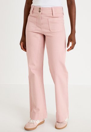 Woman wearing light pink high-waist straight-leg pants with front patch pockets and beige sneakers, standing against a white background.