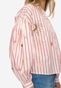 Hvid bluse med røde vertikale striber, puffede ærmer og broderede sol- og månemotiver. Har knaplukning og en afslappet pasform.