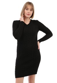 Donna con capelli biondi corti che indossa un vestito nero a maniche lunghe con scollo a V in maglia a trecce, mentre posa con una mano sul collo e l'altra sui fianchi.