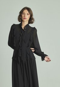 Blusa negra con volantes, mangas largas y un lazo en la cintura. Presenta un cuello alto con botones decorativos. Tejido suave y fluido.