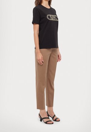 Frau trägt ein schwarzes kurzärmeliges Shirt mit Logo, beige gerade Hose und schwarze flache Sandalen, steht seitlich vor weißem Hintergrund.
