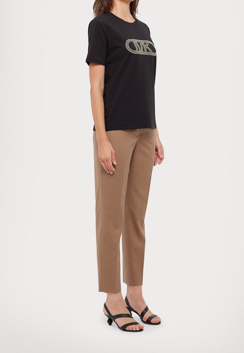 Femme portant un t-shirt noir à manches courtes avec logo, pantalon beige droit et sandales noires à talons bas, debout de profil sur fond blanc.
