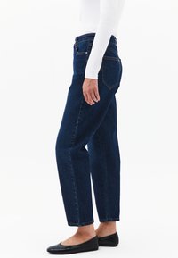 Jeans de mezclilla azul oscuro con corte recto, que presentan una cintura alta, bolsillos traseros y detalles de costura sutiles. Se llevan con zapatos planos negros.