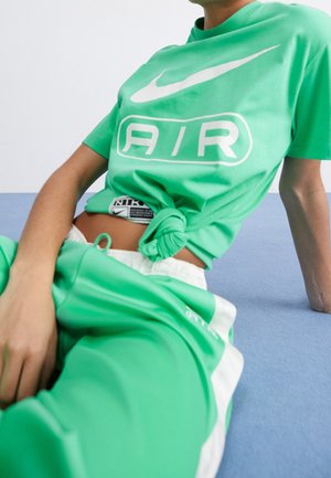 Světlá zelená tričko s krátkým rukávem a bílým logem "AIR", spárované se zelenými a bílými tepláky. Tričko uvázané v pase, což ukazuje texturu látky.