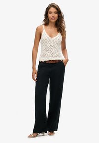 Witte gebreide tanktop met een ruitpatroon, gecombineerd met losse zwarte broek en een geweven bruine riem, aangevuld met beige sandalen.