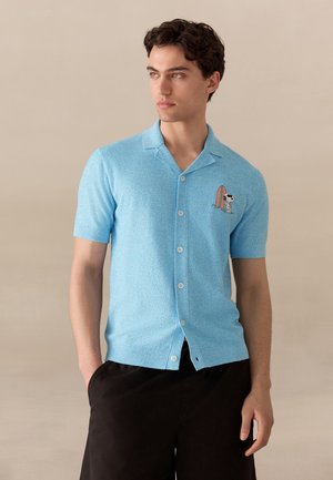 Joven con una camisa azul claro de manga corta abotonada con un parche de Snoopy y tabla de surf en el pecho, combinada con pantalones cortos negros.