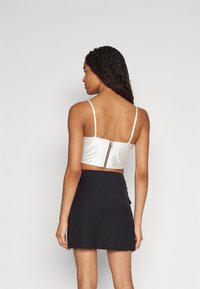 Bijela satenska crop top majica s tankim naramenicama i vidljivim zlatnim patentnim zatvaračem na leđima, u kombinaciji s crnom mini suknjom. Glatka tekstura, prilagodljiv dizajn.