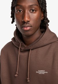 Brauner Kapuzenpullover mit Kordelzug, mit einem weißen Logo-Text "ELEMENT SKATEBOARD CO. WORLD WIDE" auf der Brust. Weicher Stoff.