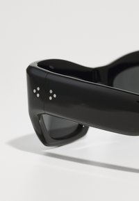 Occhiali da sole neri con un design elegante, dotati di una montatura in plastica lucida e accenti a punto argentato sulle tempie. Lenti scure.