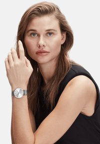 Zilveren horloge met een ronde wijzerplaat, meshband, minimale uurmarkeringen en een gladde afwerking. Model draagt een zwarte tanktop.