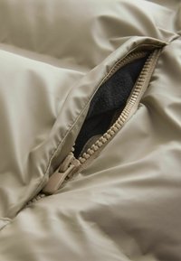 Beżowa kurtka puchowa z materiału, częściowo rozpięta, ukazując ciemne wnętrze i beżowy zamek z widocznymi zębami.