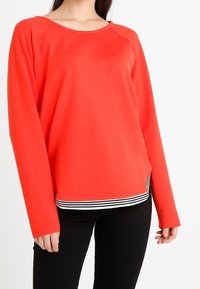 Sudadera roja de manga larga con cuello redondeado y bajo a rayas en blanco y negro. Textura suave y corte relajado.