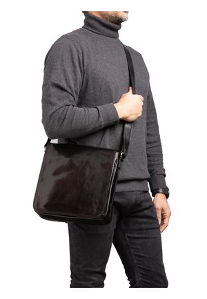Uomo che indossa un dolcevita grigio scuro e jeans neri tiene una borsa a tracolla in pelle nera con chiusura a patta.