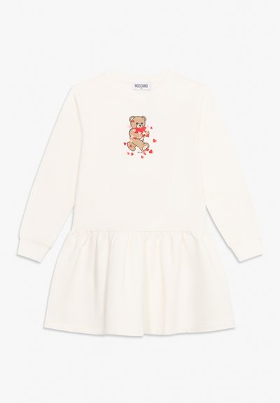 Robe blanche à manches longues avec jupe froncée, avec un ours en peluche tenant des cœurs rouges sur la poitrine et une étiquette Moschino au col.