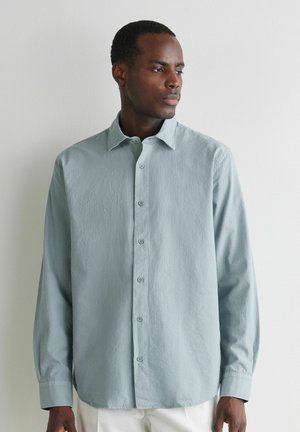 Mann trägt hellblaues, langärmliges Hemd mit Knöpfen, steht vor einer weißen Wand und schaut nach rechts.