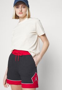 Camiseta blanca de algodón combinada con pantalones cortos deportivos negros que presentan una cinturilla roja, cordón ajustable y un logo en el bolsillo.