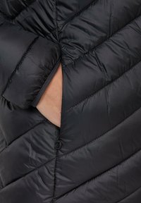 Zwarte pufferjack met diagonale gestikte naden, een zijzak en een zachte binnenvoering die zichtbaar is bij de opening van de zak.