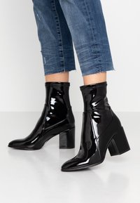 Bottines en cuir verni noir avec un bout pointu et un talon carré épais. Texture lisse et design sans coutures, légèrement au-dessus de la cheville.