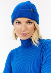 Blauer Strickmütze mit Rippenstruktur und einem Logo-Patch, kombiniert mit einem passenden, gerippten Rollkragenpullover. Beide Teile sind in leuchtendem Blau.