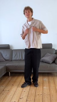 Camisa de mangas curtas, cinza claro e riscada, t-shirt branca por baixo, calças largas pretas e sapatos escuros em um chão de madeira.