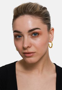Goldene Creolenohrringe mit glatter, metallischer Oberfläche. Das Model hat helle Haut, dunkles, zurückgebundenes Haar und trägt ein schwarzes, geripptes Oberteil.
