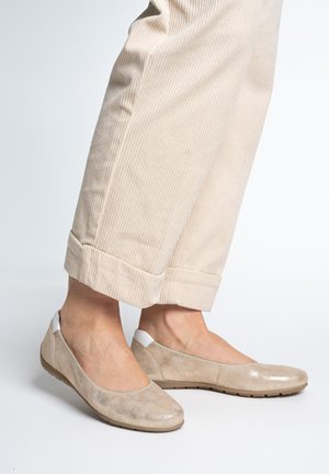 Beine mit beigen Cordhosen, die am Saum hochgekrempelt sind, und beigen flachen Slip-on-Schuhen mit weißen Fersenakzenten.