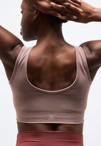 Γυναίκα με σκουρόχρωμο δέρμα που φοράει μωβ αθλητικό σουτιέν και σκουρόχρωμο πορτοκαλί κάτω μέρος, τεντώνει τα χέρια πίσω από το κεφάλι της, δείχνοντας τους μυς της άνω πλάτης.