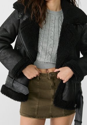 Vrouw draagt zwarte shearling jas over een grijze kabelgebreide cropped trui en een olijfgroene denim minirok, houdt de jas open.