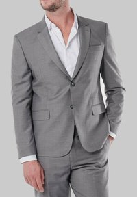 Un costume gris en tissu au toucher lisse. Il présente une fermeture à deux boutons, deux poches avant et un col cranté. Une chemise blanche en dessous.