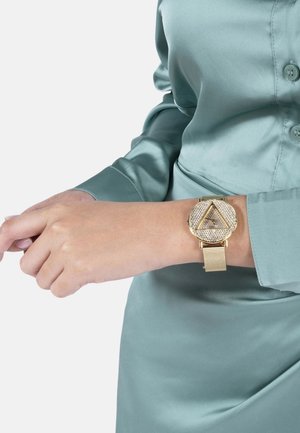 Orologio con cinturino in maglia dorata, caratterizzato da un grande quadrante triangolare decorato con dettagli simili a diamanti, indossato su un polso di pelle beige.