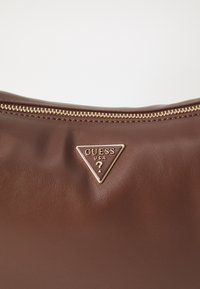 Bruine leren handtas met een gestructureerde afwerking, voorzien van een gouden rits en een driehoekige logopatch met de tekst "GUESS USA."