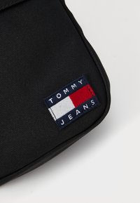 Svart tekstilveske med et rektangulært broderi med "TOMMY JEANS" tekst og horisontale striper i rødt, hvitt og blått.
