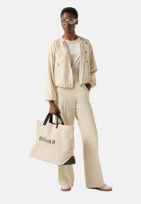 Crèmekleurige bomberjack, nauwsluitende crèmekleurige top, wijde broekspijpen en een grote canvas tas met "BOGNER" in het zwart gedrukt. Ronde lichtgekleurde zonnebril.