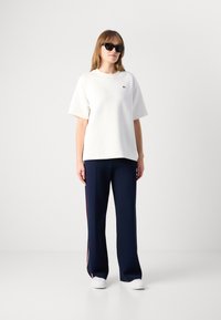 White oversized t-shirt with a small green logo, paired with navy pants featuring red side stripes and white sneakers.