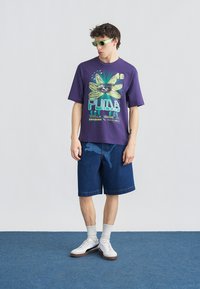 Νέος άνδρας που φορά πράσινα γυαλιά ηλίου, μοβ μπλούζα με γραφικό στοιχείο της Puma, μπλε τζιν σορτς με λογότυπο Puma, λευκές κάλτσες και λευκά αθλητικά παπούτσια, στέκεται σε μπλε χαλί.