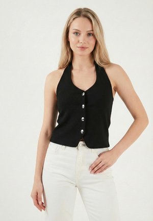 Žena v čiernom bezrukávovom halter top s gombíkmi a bielych nohaviciach, stojaca s jednou rukou na bokoch pred jednoduchým pozadím.