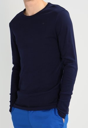 Homme portant une chemise ajustée à manches longues bleu marine et un pantalon bleu vif, debout les mains dans les poches devant un fond uni.