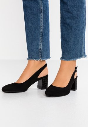 Musta mokkainen slingback-kiilakorkokenkä, jossa on kuvioitu korko, käytetty kuluneen reunan sinisten farkkujen kanssa paljain jaloin.