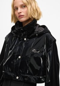 Melna īsā jaka no spīdīga materiāla ar kapuci, ar sudraba spiedpogām un izšūtu logotipu uz krūtīm. Gluda tekstūra.