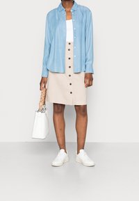 Chemise boutonnée bleu clair, débardeur blanc, jupe beige avec boutons marron, sac à main blanc et baskets blanches. Tissu lisse, coupe décontractée.