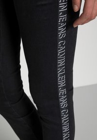 Los jeans de mezclilla negra presentan una franja lateral con "CALVIN KLEIN JEANS" en letras blancas, un diseño ajustado con textura suave y un detalle mínimo.