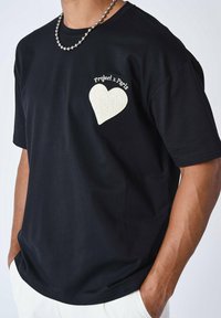 Camiseta negra de manga corta con un parche de corazón blanco texturizado y bordado "Project x Paris" en el lado izquierdo del pecho. Cuello redondo, corte relajado.