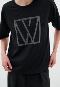 T-shirt noir à manches courtes avec un logo géométrique orné de strass. Le tissu est lisse, avec une coupe décontractée et un décolleté arrondi.