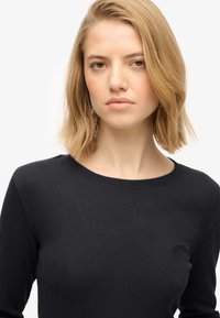 Haut noir côtelé à manches longues avec un col rond et un discret logo sur la poitrine. Texture lisse et coupe ajustée.