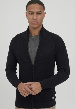 Svart strikket cardigan med høy krage og glidelås, med ribbestrikkede mansjetter og nederkant, brukt over en grå skjorte. Glatt tekstur.
