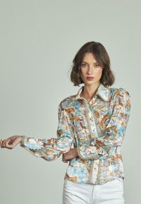 Blusa leggera a fantasia floreale in tonalità di blu soft, marrone e crema. Presenta un colletto a punta e una rifinitura decorativa lungo la parte anteriore e i polsini.