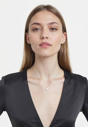 Collana d'argento con un unico pendente a perla rotonda, indossata con una maglietta nera a scollo a V, che mette in risalto il pendente su uno sfondo liscio e neutro.