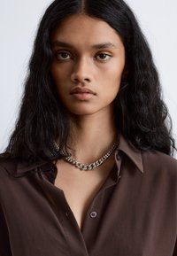Brown shirt with a collar, featuring a silver chain necklace. The shirt has a smooth texture and button detail at the chest.
