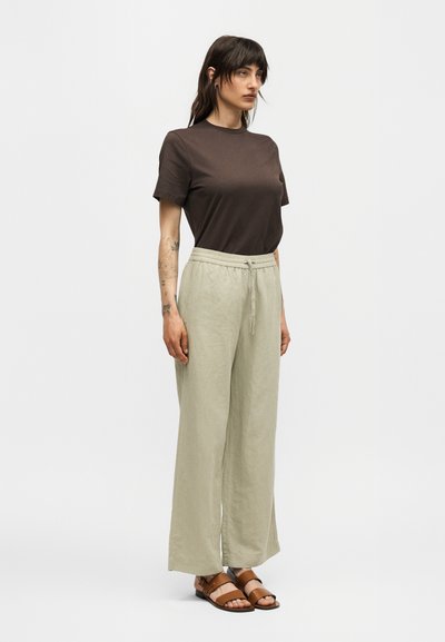 Γυναίκα με σκούρα μαλλιά, να φοράει καφέ κοντομάνικο πουκάμισο, μπεζ παντελόνια φαρδιά με κορδόνι στη μέση και καφέ σανδάλια, στέκεται μπροστά σε λευκό φόντο.