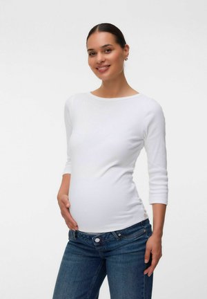 Gravid kvinde i en hvid langærmet skjorte og blå jeans smilende, med den ene hånd hvilende på maven, mod en ensfarvet baggrund.